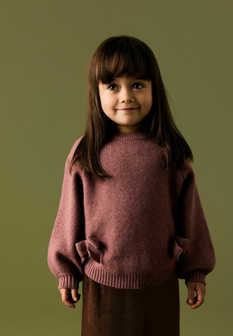 Jeune fille aux longs cheveux bruns portant un pull mauve avec des détails en forme de nœud et une jupe marron, debout devant un fond olive.