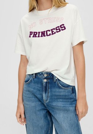 Kobieta ubrana w białą koszulkę z napisem "NO STRESS PRINCESS" w kolorze fioletowym i jasnoczerwonym, w połączeniu z niebieskimi dżinsami z wysokim stanem.