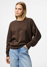 Sudadera marrón de gran tamaño con cuello redondo y mangas largas, combinada con jeans de color azul claro, que presentan un ajuste relajado y una textura suave.