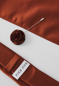 Tejido marrón acanalado con un pin de roseta decorativa y un imperdible. El tejido tiene una textura brillante. La etiqueta dice "PIER ONE."