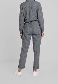 Randig jumpsuit i svart och vit med långa ärmar, elastisk midja och sidofickor; tillsammans med vita sneakers.