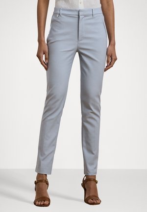 Femme portant un pantalon slim bleu clair et des talons ouverts marron, debout devant un fond clair uni.