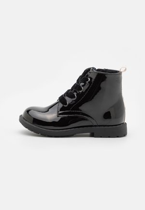 Bota de tornozelo em verniz preto com bico arredondado, atacadores pretos e uma alça na parte traseira. Sola de borracha plana com padrão texturizado.