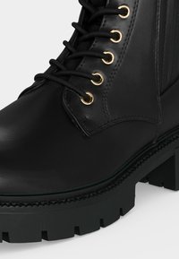 Botte en cuir noir à lacets avec œillets dorés et semelle épaisse à crampons, montrée en gros plan sur un fond clair.