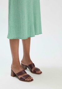 Braune Leder-Blockabsatzmules mit zwei breiten Riemen. Der Blockabsatz hat eine mittlere Höhe und der Schuh hat eine glatte Oberfläche. Darunter ein gerüschtes grünes Kleid.