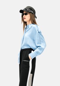 Lichtblauw satijnen shirt met een knoopsluiting, met lange mouwen en een relaxte pasvorm; gecombineerd met zwarte joggingbroek en een zwarte pet.