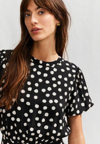 Svart blus med vita prickar, korta puffärmar, rund halsringning och mjuk textur. Har elastisk midja för en figurnära look.