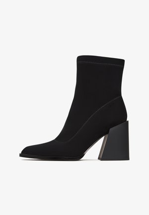 Black ankle boot made of soft fabric with a pointed toe, high block heel, and contrasting stitching details. Sleek and minimalist design.