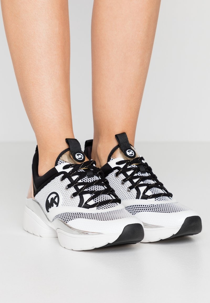 Pieds portant des baskets Michael Kors blanches et noires en tissu mesh, lacets noirs et semelle blanche épaisse sur un fond clair uni.