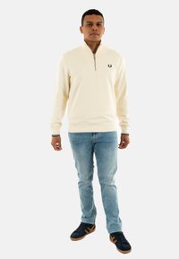 Suéter cremoso con cremallera y cuello alto, puños y dobladillo acanalados, logo negro en el pecho, combinado con jeans azul claro y zapatillas negras.