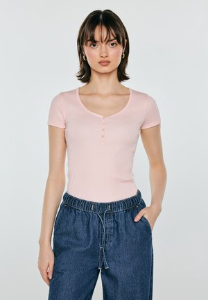 Młoda kobieta z krótkimi ciemnymi włosami, ubrana w dopasowany jasnoróżowy top z krótkim rękawem i luźne ciemnoniebieskie spodnie dżinsowe, stoi z jedną ręką w kieszeni.