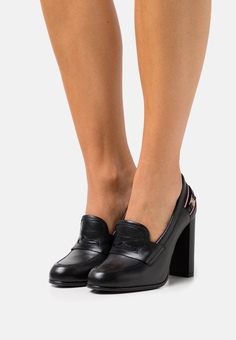 Mocassins à talons hauts en cuir noir, portés pieds nus, avec un bout arrondi, des détails cousus et un emblème décoratif près du talon.