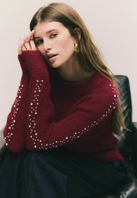 Burgundy stickad tröja med långa ärmar prydd med dekorativa rhinestones längs armen. Djup neckline och mjuk textur. Avslappnad stil.