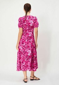 Keskikokoinen, puff-hihallinen mekko kirkkaan vaaleanpunaisena, jossa on kukkakuosi. Liukas kangas, virtaava siluetti ja takana napitusyksityiskohta. Mustat sandaalit.