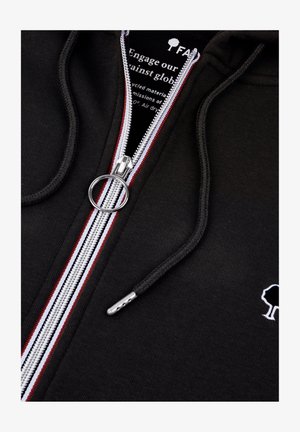 Felpa nera con zip, con strisce di accento bianche e rosse, hardware della zip in argento e un piccolo logo a forma di albero bianco sul petto. Tessuto morbido.