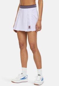 Weiße, plissierte Tennisschürze mit einem marineblauen und roten gestreiften Bund sowie einem Logo-Patch. Kombiniert mit weißen und blauen Turnschuhen sowie weißen Socken.