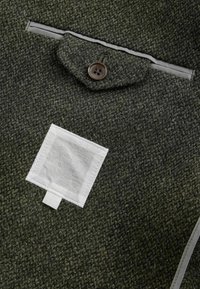Dunkelgrüner Wollstoff mit strukturierter Oberfläche, versehen mit einem Knopfverschluss und einem weißen quadratischen Etikett, das auf das Material genäht ist.