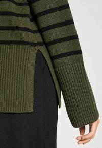 Pull tricot rayé vert et noir avec ourlet et poignets côtelés, porté avec une jupe ou un pantalon noir, main détendue sur le côté.