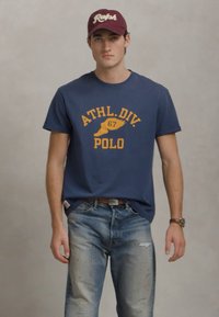 Mladý muž v námořnickém tričku s žlutým nápisem "ATHL. DIV. POLO", opraných džínách, vínové čepici s bílým nápisem "Ralph" a s hodinkami na zápěstí.