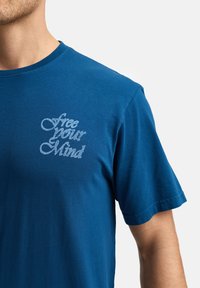 Camiseta de algodón azul royal con mangas cortas y cuello redondo. Texto "libera tu mente" en una escritura azul más clara en el área del pecho izquierdo.