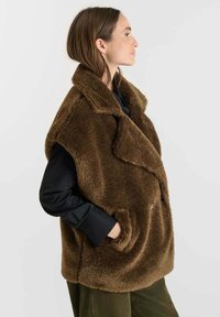 Nepbont gilet in donkerbruin met een sjawlkragen, oversized ontwerp, zijzakken en een gestructureerd afwerking. Draagt over een zwart shirt.