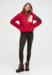 Rode pufferjack met een gewatteerd ontwerp, lichtroze borstzak en blauwe mouwpatch. Draag het met olijfgroene legging en bruine wandelschoenen.