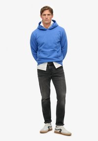 Felpa blu con tasca a canguro, indossata sopra una maglietta bianca, abbinata a jeans scuri e sneakers bianche con dettagli neri.