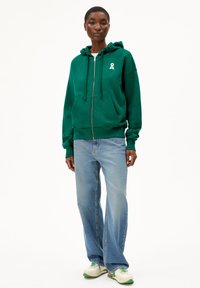 Grüne Zip-Hoodie mit Kordelzug, versehen mit einem kleinen weißen Band-Logo. Getragen mit hellblauen Schlagjeans und mehrfarbigen Turnschuhen.