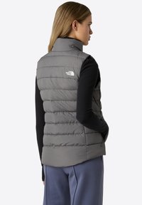 Harmaa puffervesti, jossa on korkea kaulus ja vaakasuora tikkauskuvio. Varustettu logolla yläselässä ja sivuvetoketjulla.