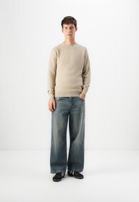Giovane uomo in piedi con le mani nelle tasche, indossando un maglione beige, jeans a gamba larga blu e sneakers nere su sfondo bianco.