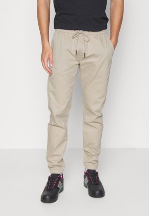 Beige Jogginghosen mit Kordelzug, getragen von einer Person mit schwarzem Hemd und schwarzen Sneakers mit rosa und grünen Akzenten.
