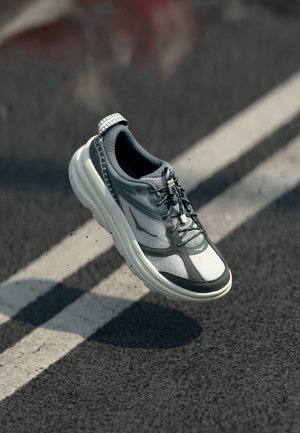 Baskets grises et blanches avec lacets en l'air au-dessus d'une route asphaltée marquée de lignes blanches, projetant une ombre en dessous.