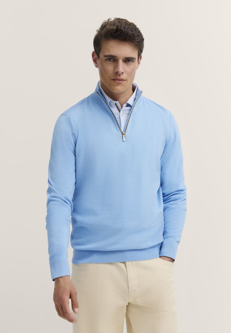 Hellblaues Strickpullover mit einem halben Reißverschluss-Kragen, gerippten Bündchen und Saum. Getragen über einem weiß-blau gestreiften Hemd. Glatte Textur.