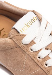 Zapatilla de ante marrón claro con cordones blancos, ojetes y una etiqueta blanca con letras doradas en la lengüeta; punta redondeada y superficie texturizada.