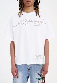 Hvid bomulds t-shirt med korte ærmer, der har "Ed Hardy" i lysegrå kursiv skrift og et lille logo ved kanten.