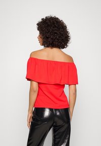 Haut rouge à épaules dénudées avec un large volant, manches courtes, texture lisse et silhouette ajustée, associé à un pantalon noir brillant.