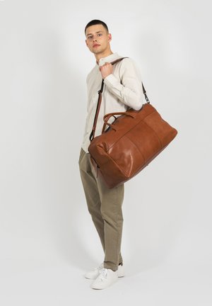 Brown leather duffel bag with two top handles, adjustable shoulder strap, and zipper closure. Strong seams and smooth texture.