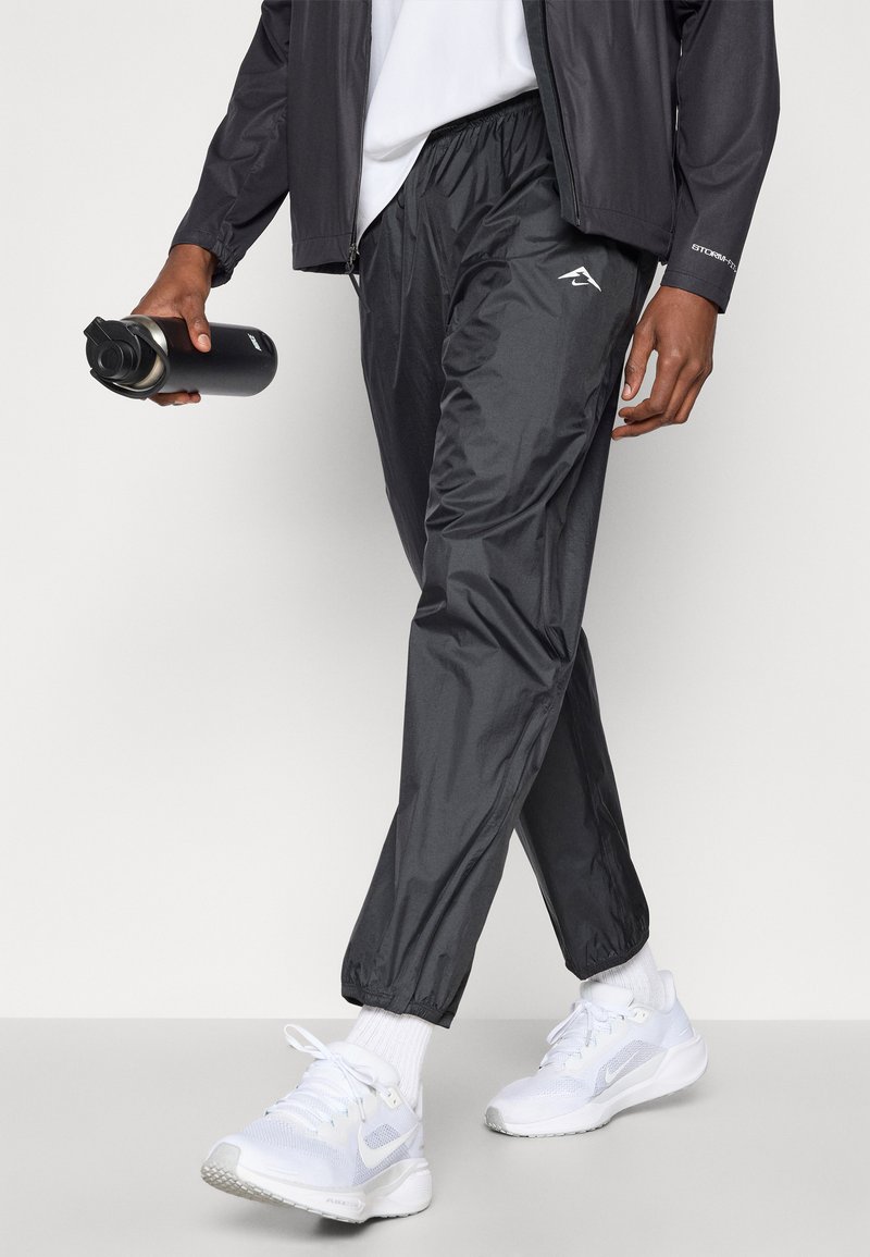 Zwarte, lichte sportbroek met een elastische taille en boorden, gecombineerd met witte sneakers en een zwarte waterfles.