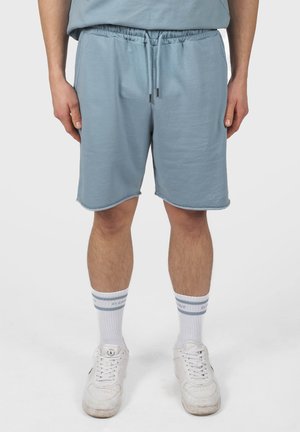 Hombre con pantalones cortos de color azul claro con cordón, calcetines blancos y zapatillas blancas, de pie frente a un fondo liso.