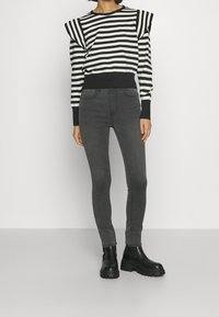 Suéter a rayas blanco y negro con hombros abullonados, combinado con jeans ajustados gris oscuro y botas de tobillo negras con suelas gruesas.