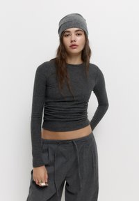 Graues langärmliges Oberteil mit tailliertem Schnitt und strukturiertem Stoff, kombiniert mit lockeren grauen Hosen. Eine passende graue Mütze rundet das Outfit ab.