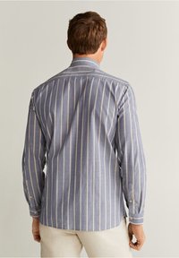 Homme portant une chemise à manches longues rayée verticalement bleu et blanc et un pantalon de couleur claire, vu de dos sur un fond uni.