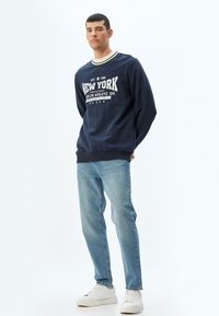 Felpa navy con testo grafico bianco, dettagli a costine sul colletto e sui polsini, abbinata a jeans azzurri chiari e sneakers bianche. Sfondo semplice.