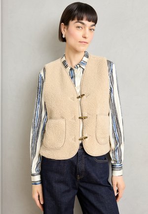 Beige fleece vest med to frontlommer, messinglukninger og trekantede detaljer. Båret over en stripete skjorte med knapper og mørke jeans.