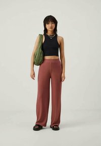 Μαύρο ριχτό crop top, καφέ μπορντώ παντελόνι με φαρδύ πόδι και υφασμάτινη πράσινη τσάντα. Απλές σανδάλια ολοκληρώνουν την εμφάνιση. Ουδέτερο φόντο.