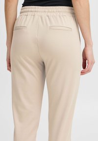 Beige elastische taillebroek van gladde stof, met twee achterzakken en een relaxte pasvorm. Eenvoudig ontwerp zonder patronen of versieringen.