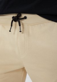 Lys beige joggerbukser lavet af blødt stof med snor i taljen, med sorte detaljer og en sidelomme. Glat tekstur med ribbet talje.