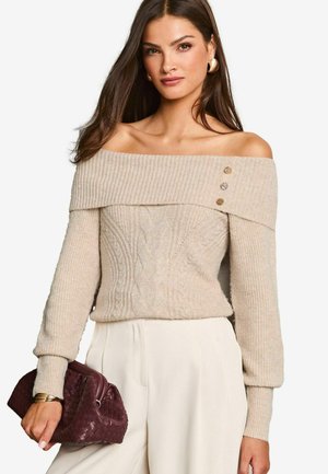 Pull beige à épaules dénudées avec un col côtelé et une texture en tricot torsadé, agrémenté de boutons décoratifs. Porté avec un pantalon blanc et un sac bordeaux.