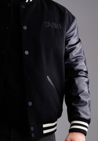 Schwarze Collegejacke mit Wollkörper und Lederärmeln, ausgestattet mit einem Druckknopfverschluss, gesticktem Logo und gestreiften Rippbündchen.