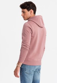 Felpa rosa chiaro con cappuccio regolabile, polsini e orlo a costine, indossata sopra un paio di jeans blu, vista da dietro.